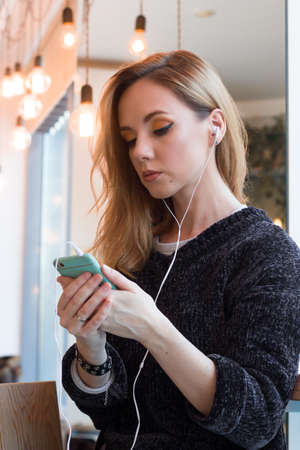 beautiful smiling young woman in gray sweater using mobile phone with earphones to listen to music,...