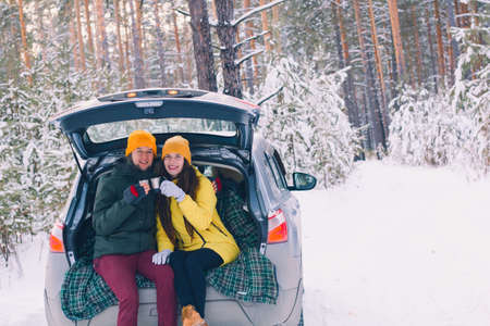cute couple in love sitting in a car with an open trunk with mugs of hot drinks in the winter forest  between  of the trees covered with snowの写真素材