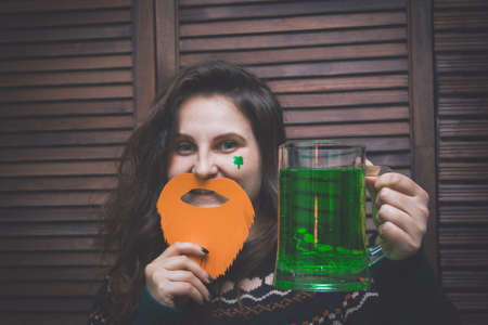 young beautiful woman with a red paper beard and a mug of green ale in a bar opposite a wooden wallの写真素材