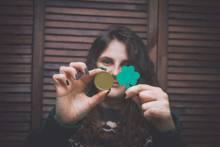 young beautiful woman with painted green clover on her cheek holding golden coin at the bar in front...