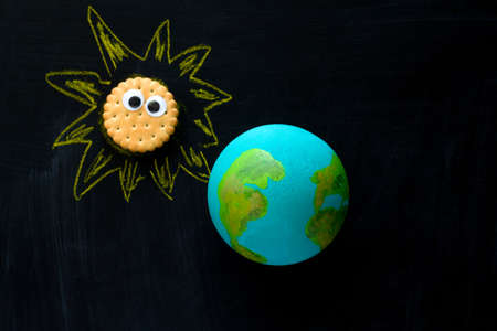 top view  handmade model  of Earth planet  and cookie Sunwith funny googly eyes  on the chalkboard ,...