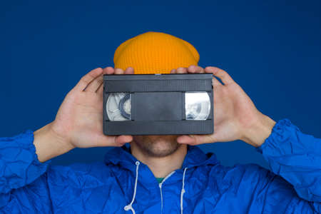 young man  in 90s sports jacket and yellow hat with VHS cassette on blue backgroundの写真素材
