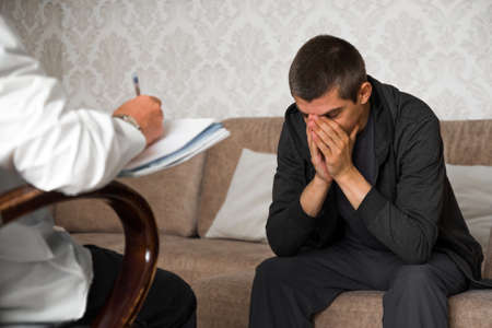 young man is sitting on couch and covers face with hands  in  the psychologist officeの写真素材