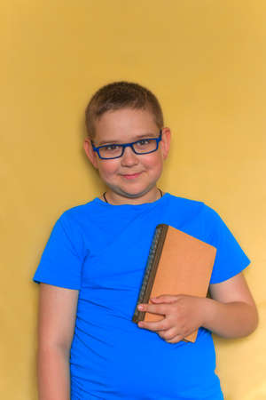 portrait of  young cheerful school boy, smiling, wearing glasses holding a book against yellow...