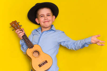 beautiful cute little boy in hat and shirt keeps guitar isolated on yellow background