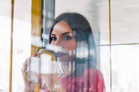 beautiful woman in red dress holding take away coffee cup in a coffee shop, shot through the glassの写真素材