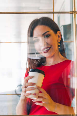 beautiful woman in red dress holding take away coffee cup in a coffee shop, shot through the glass