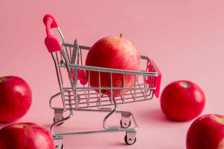 little metal shopping cart with red apples on a pastel pink background, healthy eating and autumn apple harvest conceptの写真素材