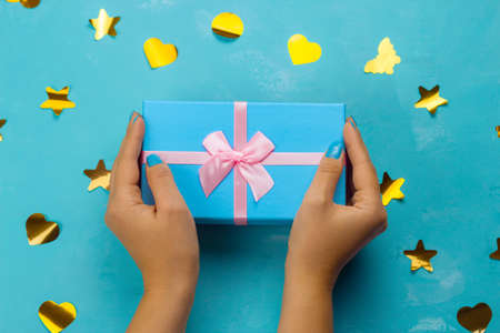 top view female hands holding blue gift box with festive pink bow on a blue background with golden confettiの写真素材