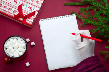 top view flat lay sheet of blank paper with space for text and Santa Claus hand in white glove , fir branches, gift box and cocoa cup on a red backgroundの写真素材