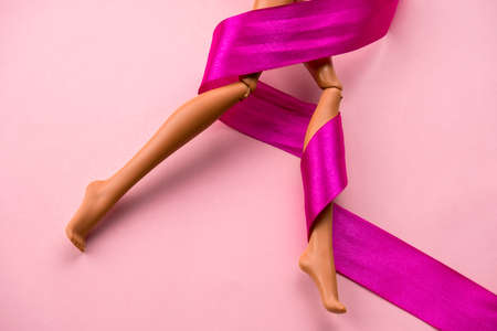 Close up of smooth legs of a plastic toy doll with pink satin ribbon  on a soft pink background.の写真素材