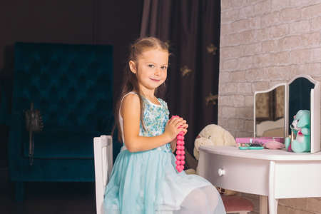 Little caucasian girl in a blue dress sits on a chair at the dressing table and holds pink beads in her handsの写真素材