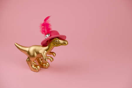 funny golden dinosaur toy wearing pink hat with feather on a soft pink backgroundの写真素材