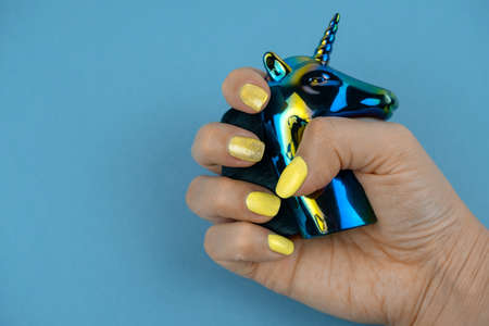 beautiful well groomed female hand with yellow manicure hold unicorn on a blua background copy space