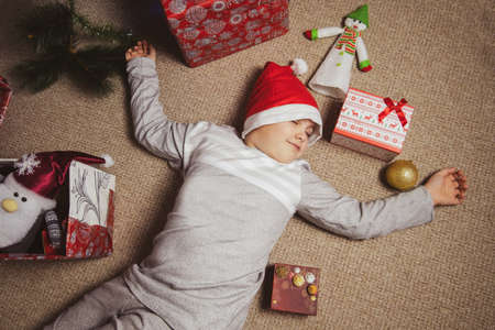 cute little boy in gray pajamas and Santa hat sleeps on the floor under a Christmas tree among...