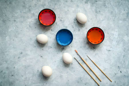 top view selective focus cups with dye, brushes and white eggs on a concrete background, painting...