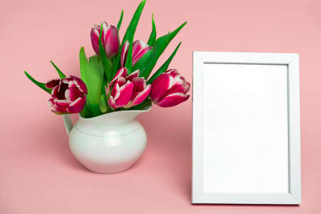 beautiful fresh pink tulips in a white porcelain jug and white wooden mock up photo frame on a...