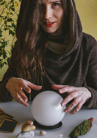 beautiful young mysterious woman in a hoodie holds her hands over a crystal ball of predictions and...
