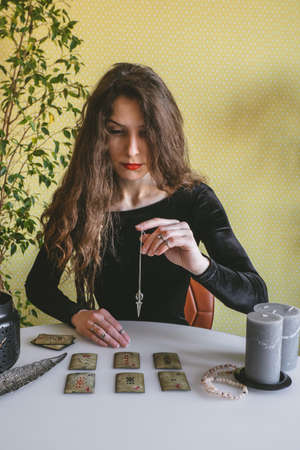 beautiful young woman in a black velvet dress holds a crystal on a silver chain above the runes during a spiritual seanceの写真素材