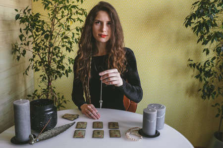 beautiful young woman in a black velvet dress holds a crystal on a silver chain above the runes during a spiritual seanceの写真素材
