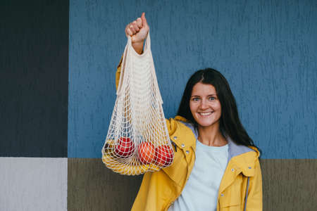 young happy woman holding net shopping bag on the street against gray wall, copy space, emotional female portraitの写真素材