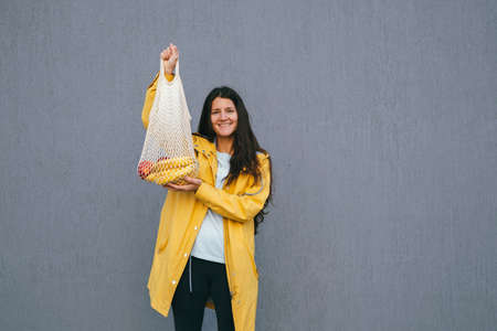 young happy woman holding net shopping bag on the street against gray wall, copy space, emotional female portraitの写真素材