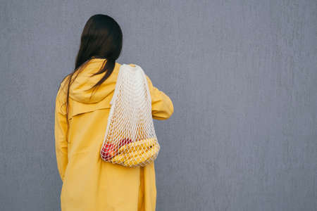 young woman with long dark hair wearing yellow raincoat holding string shopper bag with fruits standing back to the camera against gray wallの写真素材
