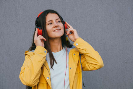 pretty young brunette woman wearing yellow raincoat and using red headphones listening music during walking outdoor against gray wall, lifestyle portrait copy spaceの写真素材