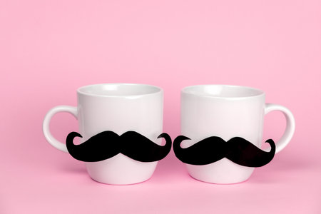 White ceramic cups decorated with black mustache. Cute romantic card for gay couple on Valentine's day on a pink background.の写真素材