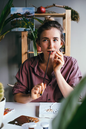 Young beautiful woman is sitting at work desk and holding a brush near her face. Searching ideas and inspiration concept.の写真素材
