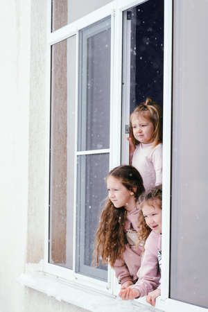 Three little girls lean out of the window and look at the falling snowの写真素材