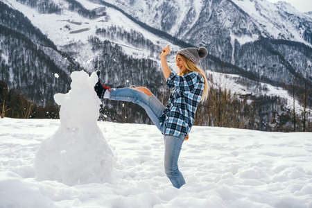 Young woman dressed in casual clothes kicks a snowman against the backdrop of mountains. Winter fun concept.の写真素材