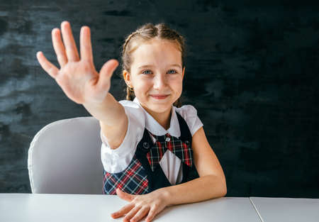 Cute schoolgirl sitting at her desk with her hand raised against the background of a black chalkboard.の写真素材