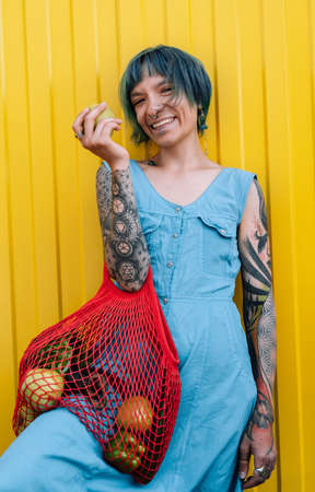 Happy beautiful woman in blue dress holds apple and shopper with green vegetables against yellow wall. Vegetarianism concept.の写真素材