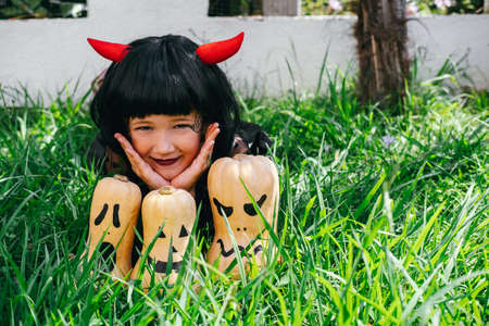 Cheerful little girl in black wig with horns and black dress sits on grass among pumpkins. Autumn,...