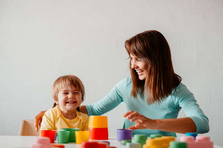 Young woman plays with her little daughter. Caucasian woman and child are sitting at table and playing against white wall. Happy family together.の写真素材