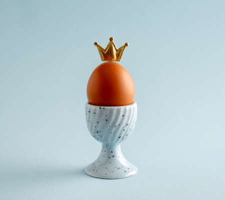 Cute brown egg wearing tiny golden crown , standing on egg stand. blue pastel background. Easter and leadership concept.の写真素材
