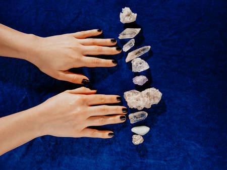 Top view female hands with black nails lying on table with gem stones. Blue velvet background.