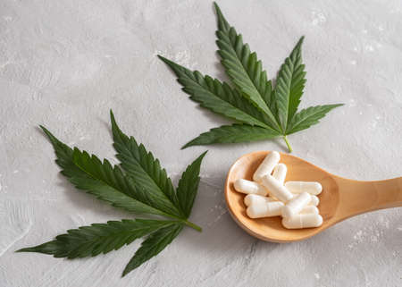 Spoon full of white capsules and green cannabis leaves on gray concrete background.の写真素材