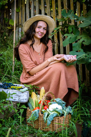 Beautiful caucasian woman wearing straw hat and linen dress sitting in garden with baskets full of fruits and vegetables crop. Autumn harvest and vegetarian organic harvest crop.の写真素材