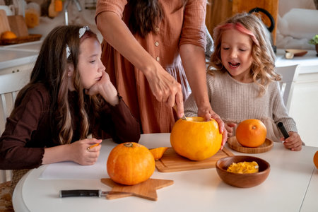 Mother holding knife ang carving spooky face on pumpkin in light white kitchen at home with her happy little daughters. Halloween party preparation.の写真素材