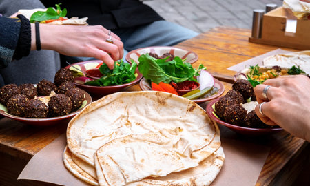 Close-up of female and male hands holding bread and spreading hummus. A plate of falafel and vegetables on the table. middle east cuisine.の写真素材
