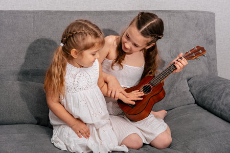 Two girls are sitting on the couch with a ukulele. The older sister teaches the younger to play the guitar.の写真素材
