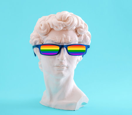 Plaster statue head wearing glasses in colors of rainbow flag on blue background.の写真素材