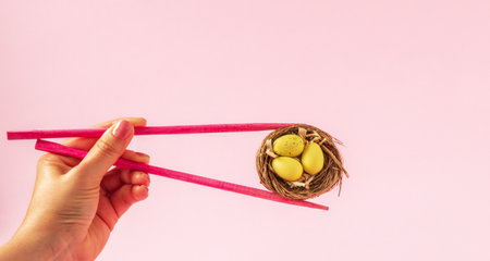 Female hand holding chopsticks and little nest with painted Easter eggs on pastel pink background. Sushi shop Easter spring advertisment concept.の写真素材