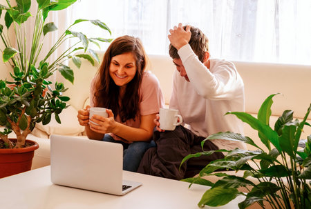 Young couple of man and woman are laughing while watching a video on a laptop sitting on the couch in their bright apartments.の写真素材