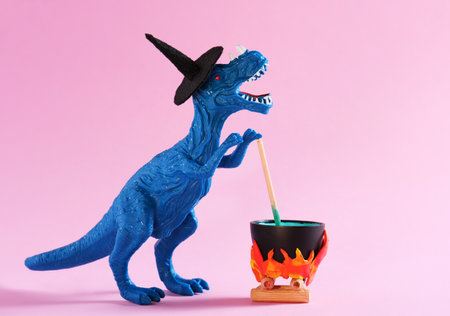 Blue dinosaur wearing witch hat is brewing potion in pot on pink background. Halloween greeting card.の写真素材