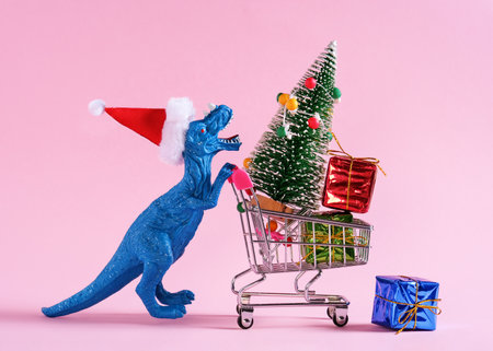 Happy blue dinosaur in Santa Claus hat carrying shopping cart with Christmas tree and gift boxes on pastel pink background.の写真素材