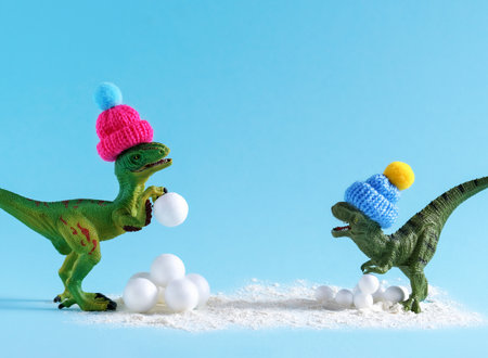 Cute green dinosaur in knitted hats playing snowballs. Happy winter holidays card.の写真素材