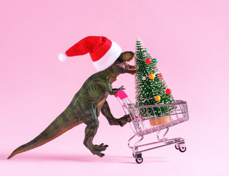 Happy green toy dinosaur carrying shopping cart with Christmas tree on pastel pink background. Minimal trendy art cart for winter holidays.の写真素材